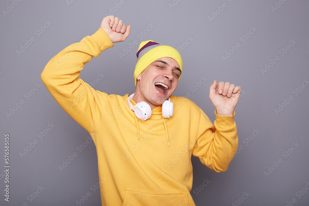 Photo of extremely happy overjoyed hipster man wearing yellow hoodie, beanie hat headphones standing isolated over gray background, rejoicing, celebrating, dancing with raised arms. Photo of extremely happy overjoyed hipster man wearing yellow hoodie, beanie hat headphones standing isolated over gray background, rejoicing, celebrating, dancing with raised arms.
