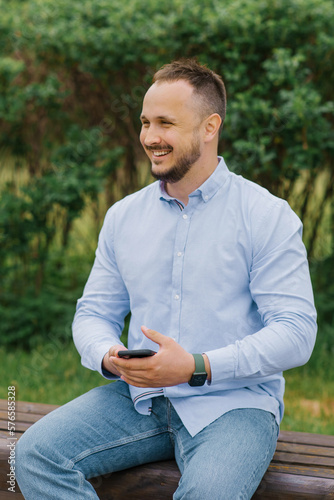 Smiling man holds a smartphone in his hands outdoors, sitting on a bench