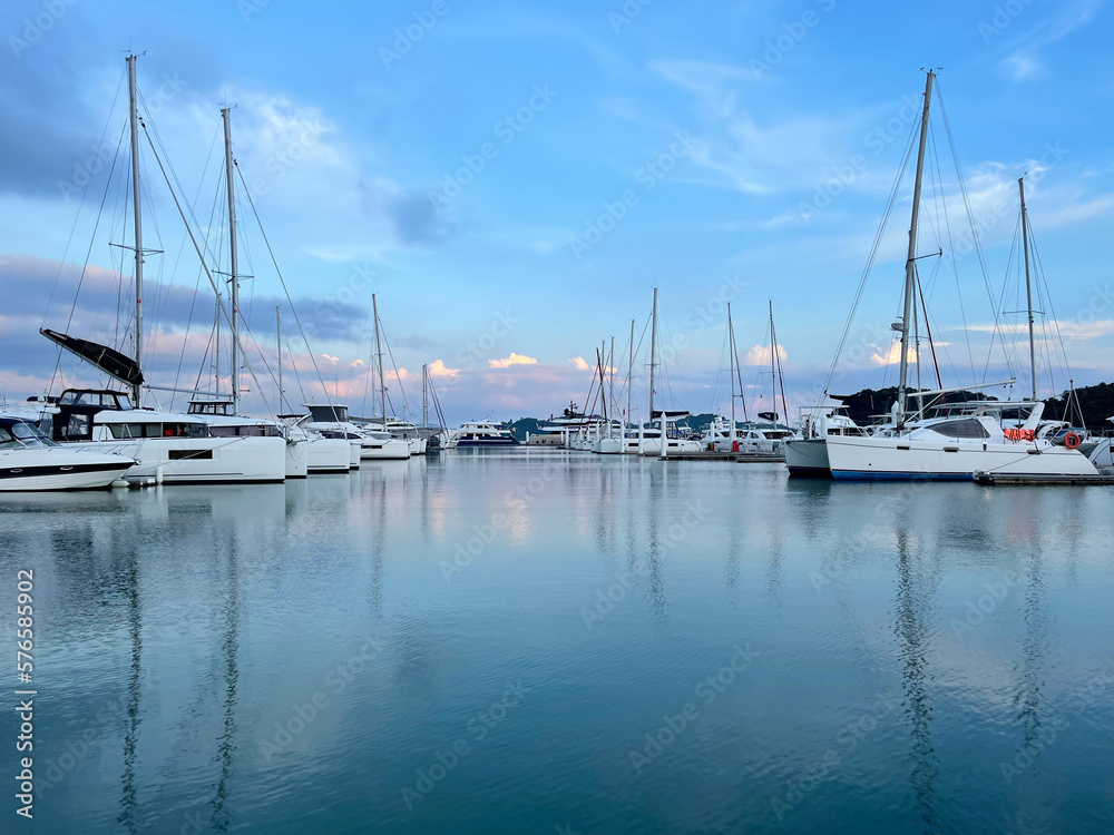 Boats in marina. Beautiful yachts in the bay at sunset are moored in a row. Amazing sky with pink glow and clouds. Sailboats at anchor. Sea harbor. Luxury yacht club. Phuket island, Thailand.