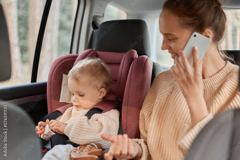 Portrait of beautiful young mother wearing beige sweater talking on the mobile phone in the car while the child is sitting next to her in baby car seat, traveling by automobile. Portrait of beautiful young mother wearing beige sweater talking on the mobile phone in the car while the child is sitting next to her in baby car seat, traveling by automobile.