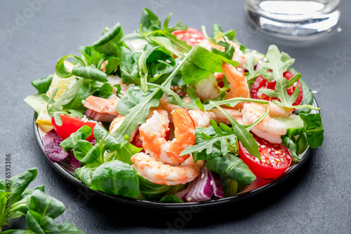 Fresh shrimp salad with cherry tomatoes, arugula, lamb lettuce, cucumber, red onion and sesame seeds on black table background. Top view