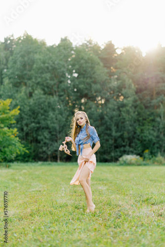 Young Caucasian woman or girl in a summer dress and a denim shirt is walking on the grass, holding shoes in her hands, relaxing outside the city on a weekend in summer