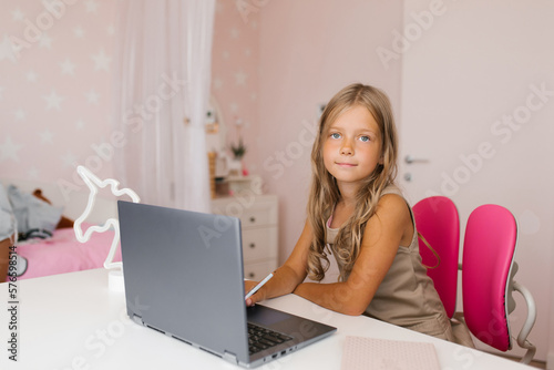 Little girl uses a video tutorial on a laptop, sitting at a table in a room and looking at the camera. Online training. Remote school for children.