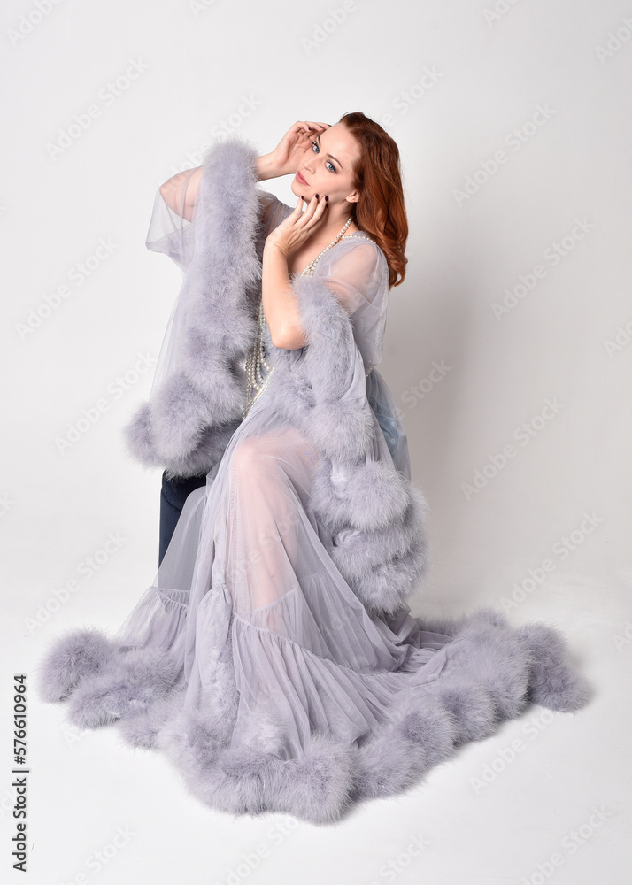 Naklejka premium Portrait of beautiful red haired woman wearing glamorous fluffy bridal dressing gown and vintage pearl necklace. Elegantly seated pose, isolated on white studio background.