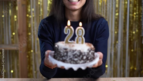 Birthday celebration concept. Woman holding birthday cake with burning candles number 28 and looking at the camera at home. Point of view or POV. Copy space photo