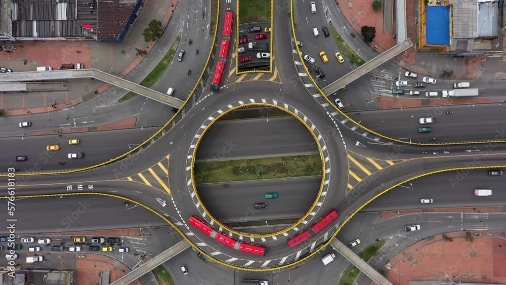 A bird's eye view shot with a 4K drone descending on a round point intersection with red articulated buses, taxis and other vehicles turning from Third street to 30th avenue in Bogotá, Colombia.