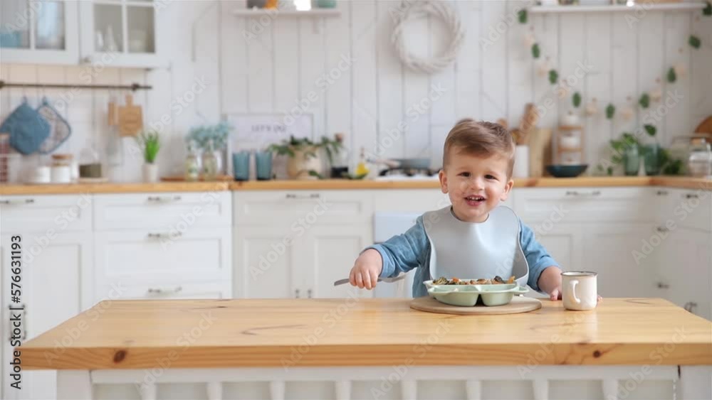 Cute caucasian toddler boy eats vegetables and meat on his own using a fork. Self-feeding concept. BLW. The child eats healthy vegetables with meat on a high chair in the kitchen. Copy space, Mock up Cute caucasian toddler boy eats vegetables and meat on his own using a fork. Self-feeding concept. BLW. The child eats healthy vegetables with meat on a high chair in the kitchen. Copy space, Mock up