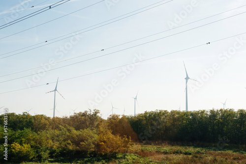 13 wind turbines producing electricity above forest with multiple high voltage electricity lines above them - green forest location
