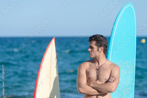 Hispanic surfer guy standing on the beach in a swimsuit looking at the camera next to two surfboards.