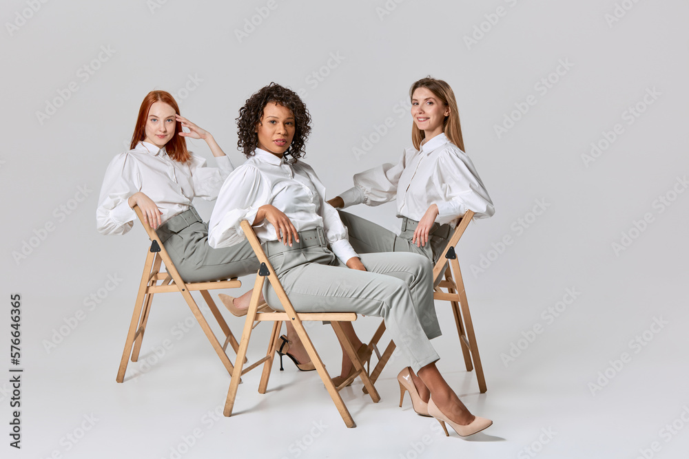 Fototapeta premium Portrait of adorable stylish young women in office style clothes sitting on chairs over light background. Concept of beauty, fashion, business, ad