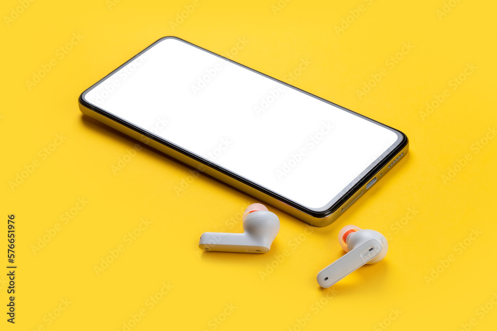 Mobile phone with empty screen and earphones on yellow surface. Wireless earphones, mobile phone on yellow background Mobile phone with empty screen and earphones on yellow surface. Wireless earphones, mobile phone on yellow background