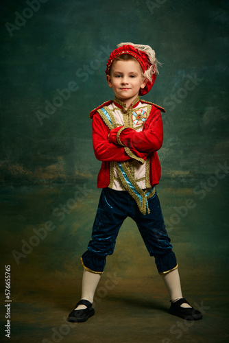 Portrait of cute little boy dressed up as medieval character, little prince and pageboy posing over dark vintage style background, Fashion, emotions, theater art concept photo