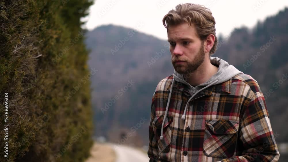 A young blond man touches a shrub thoughtfully, lost in contemplation. The serene natural surroundings provide a peaceful backdrop to this moment of introspection. A young blond man touches a shrub thoughtfully, lost in contemplation. The serene natural surroundings provide a peaceful backdrop to this moment of introspection.
