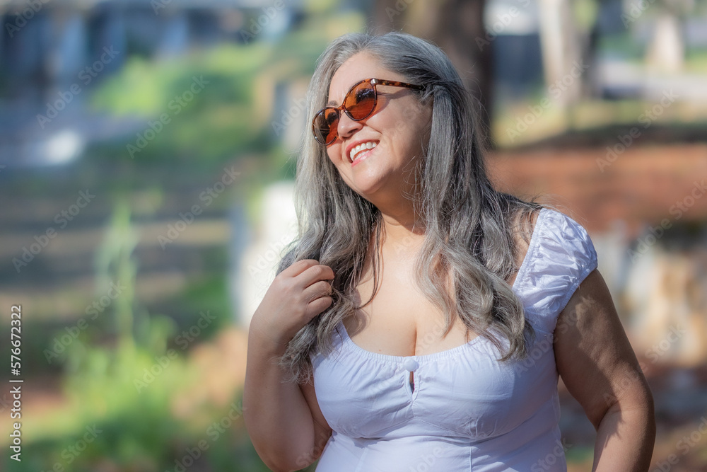 Waist up portrait of senior woman with a wide smile standing against blurred background, head turned to the side, long gray hair, sunglasses, white blouse, sunny day in the park