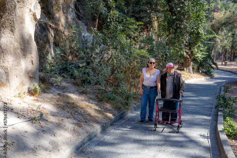 Adult daughter supporting her senior mother when taking her for walk with walker, they walk in park on a spring day, enjoying a sunny day surrounded by trees, casual clothing, sunglasses and hat Adult daughter supporting her senior mother when taking her for walk with walker, they walk in park on a spring day, enjoying a sunny day surrounded by trees, casual clothing, sunglasses and hat