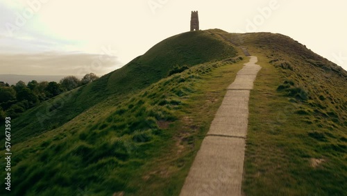 A drone shot flying along a foot path up Glastonbury Tor, a hill in Somerset UK. The shot climbs to reveal a view of green fields that stretch to the horizon photo