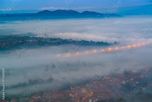 Aerial view of the town in the early morning mist is beautiful in the highlands. Low clouds and fog cover the sleeping city, photo in full moon light. Alpine mountain valley mists landscape at dawn