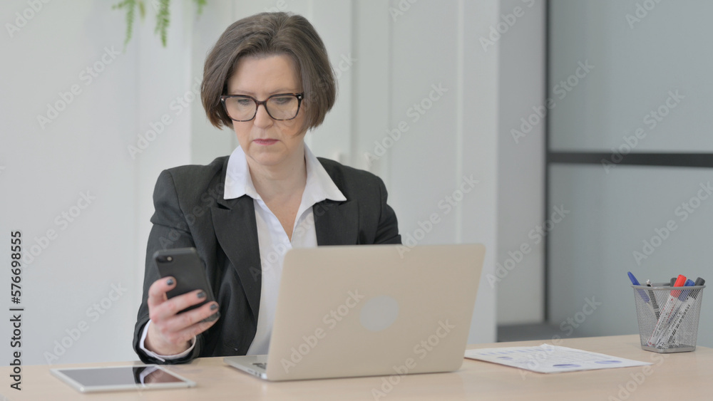 Old Businesswoman using Smartphone while using Laptop