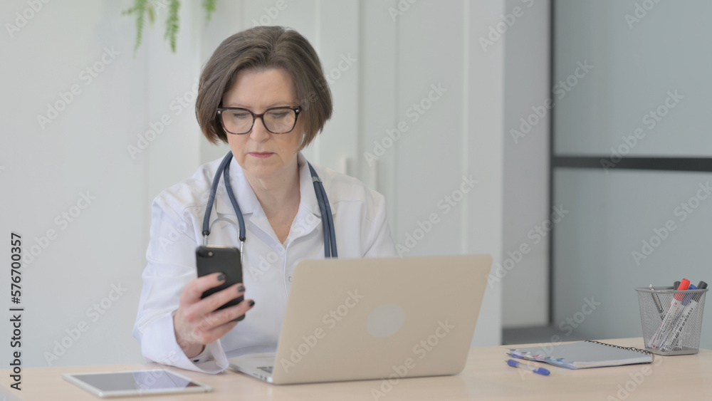 Old Female Doctor using Smartphone while using Laptop Old Female Doctor using Smartphone while using Laptop