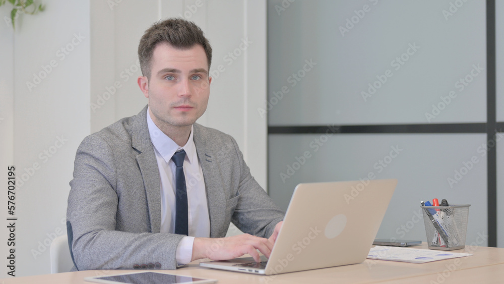 Young Businessman Looking at Camera while using Laptop Young Businessman Looking at Camera while using Laptop
