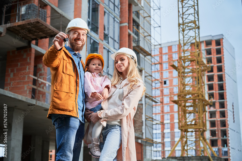 Man holding apartment keys and smiling while standing next to wife and daughter outside building under construction. Happy family homeowners posing on the street at construction site.