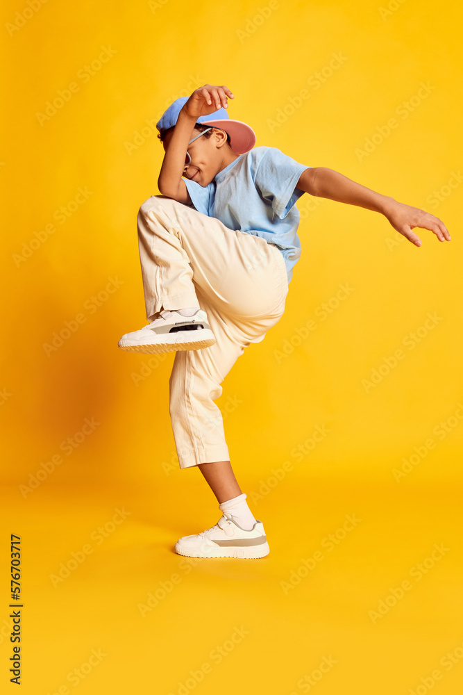 Dynamic portrait of little african boy, hip-hop dancer in stylish street style clothes dancing over bright yellow background. Concept of music, dance happiness