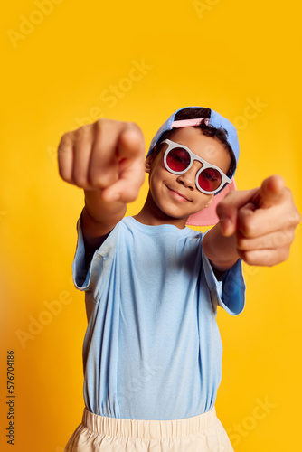Closeup portrait of little african happy boy in stylish sunglasses and cap looking at camera over bright yellow background. Concept of music, happiness, kids emotions