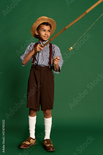 Cute handsome little boy, child in checkered shirt and suspender shorts isolated over dark green background. Concept of childhood, friendship, retro fashion style