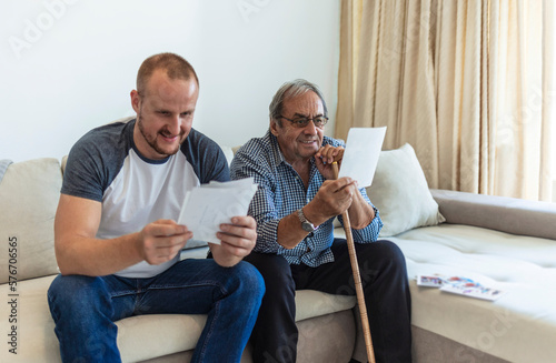 An adult hipster son and senior father sitting on stairs indoors at home, looking at photographs. Senior father and adult son talking and looking at pictures at home.