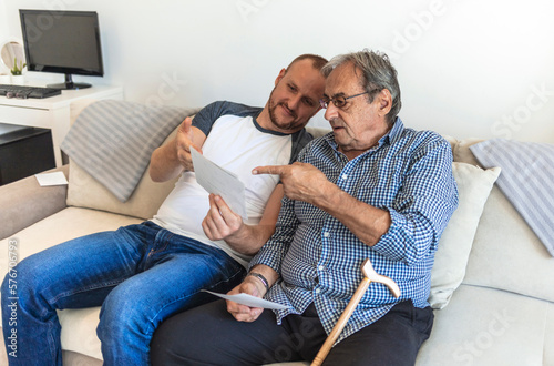 Caring loving adult son hugging embracing his old elderly senior father while he is showing his family photograph photo at home, telling stories of his youth. Happy father`s day! © Jelena Stanojkovic