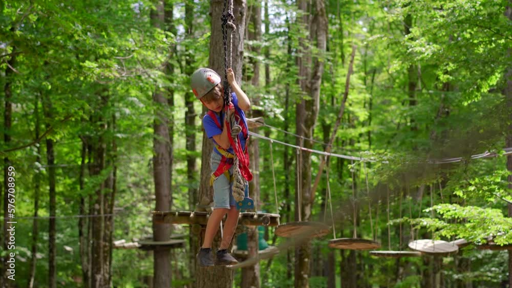 A little boy is enjoying an exciting adventure on a rope obstacle course in a lush forest. He is wearing a helmet and a safety harness as he navigates through the challenging course, showing bravery A little boy is enjoying an exciting adventure on a rope obstacle course in a lush forest. He is wearing a helmet and a safety harness as he navigates through the challenging course, showing bravery