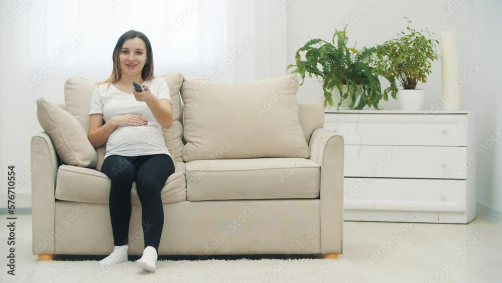 4k video of pregnant woman sitting on white sofa with remote control. 4k video of pregnant woman sitting on white sofa with remote control.