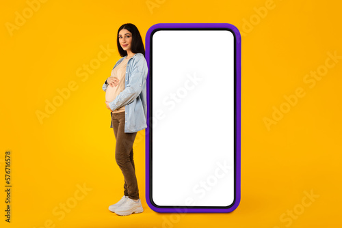 Happy pregnant woman leaning at big smartphone with blank screen and embracing her belly, advertising pregnancy app