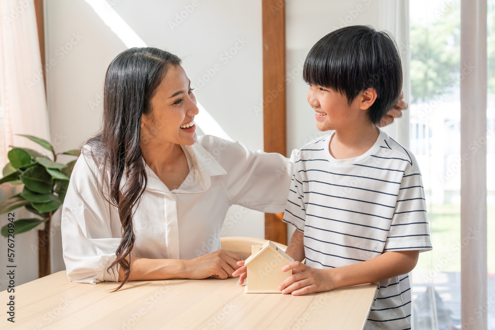 Asian mother looking her son saving money put coins into home bank model at home Asian mother looking her son saving money put coins into home bank model at home