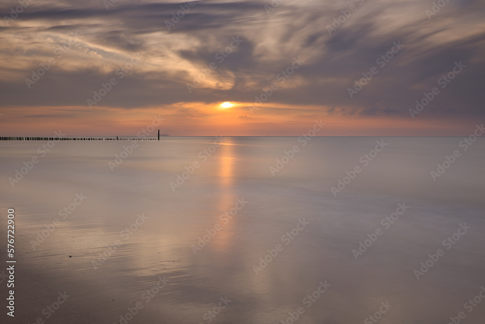 Horizontal view on row of pile heads leading into water. North sea beach during sunset with sun and clouds at the sky. Calm seascape of breakwaters in Zeeland with copy space, long exposure