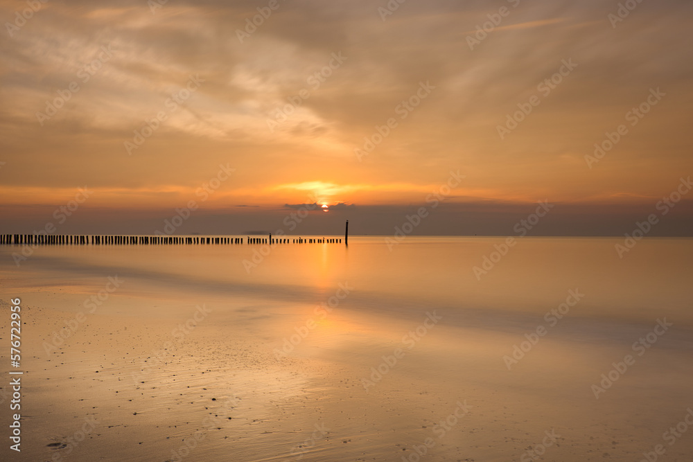 Horizontal view on row of pile heads leading into water. North sea beach during sunset with sun and clouds at an orange sky. Calm seascape of breakwaters in Zeeland with copy space, long exposure