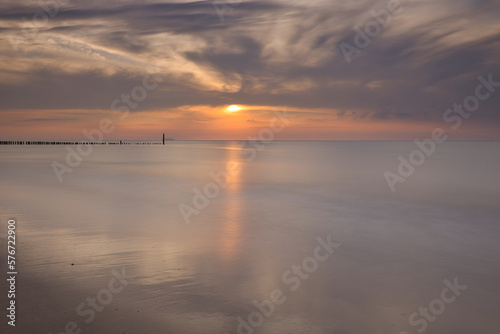 Horizontal view on row of pile heads leading into water. North sea beach during sunset with sun and clouds at the sky. Calm seascape of breakwaters in Zeeland with copy space  long exposure