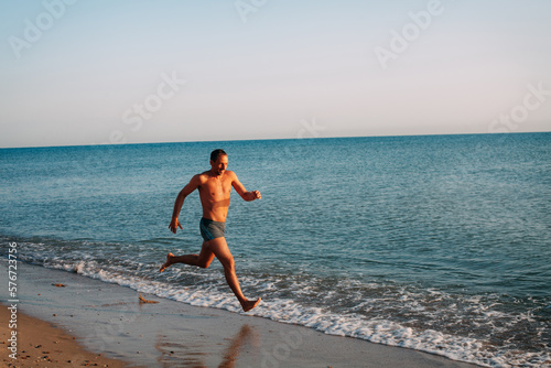 A slender muscular tanned guy in blue swimming trunks runs along the sandy shore of the ocean and smiles on a bright sunny day.