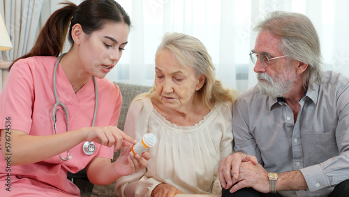 Caucasian young adult nurse is visiting elderly couple and holding a medicine bottle to explain how to use this medicine. Female caregiver is providing advice on health care to senior grandparents.
