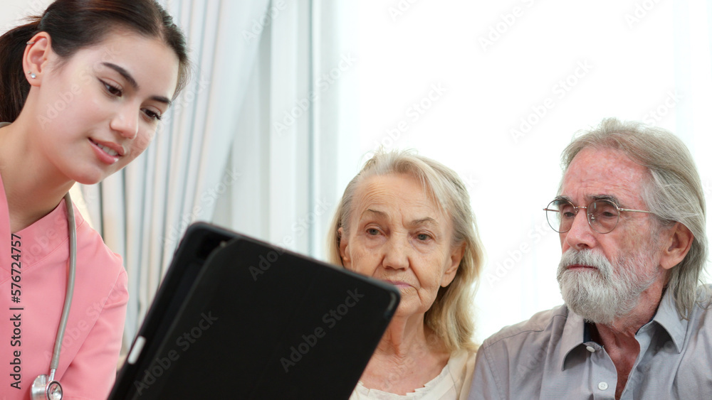 Caucasian young adult nurse is visiting elderly couple and holding a tablet to show medical records. Female caregiver is explaining result of medical examination and treatment to senior grandparents. Caucasian young adult nurse is visiting elderly couple and holding a tablet to show medical records. Female caregiver is explaining result of medical examination and treatment to senior grandparents.