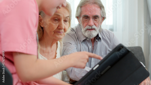 Caucasian young adult nurse is visiting elderly couple and holding a tablet to show medical records. Female caregiver is explaining result of medical examination and treatment to senior grandparents. 