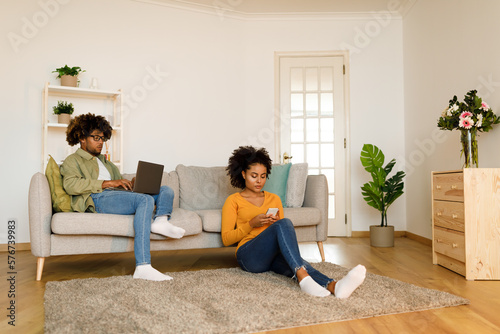 Black Couple Using Laptop And Phone Sitting In Living Room