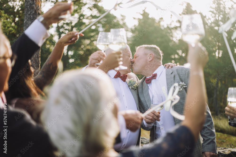 Newlywed gay couple kissing in front of guests raising toasts at