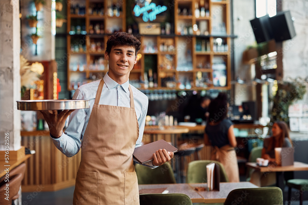 Young happy man working as waiter in cafe and looking at camera.