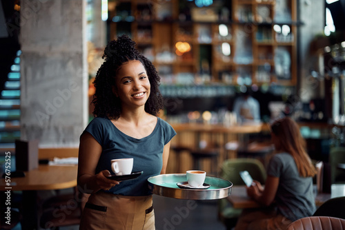 Happy black waitress serving coffee in cafe and looking at camera.