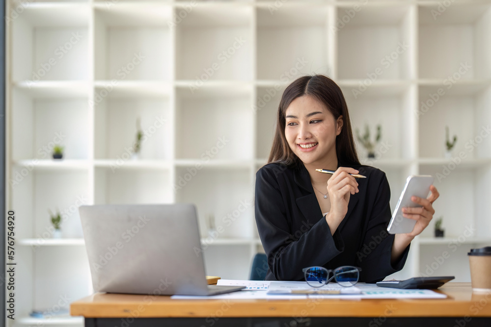 Asian businesswoman using smartphone to do accounting work with laptop and graph of financial reports in office Asian businesswoman using smartphone to do accounting work with laptop and graph of financial reports in office