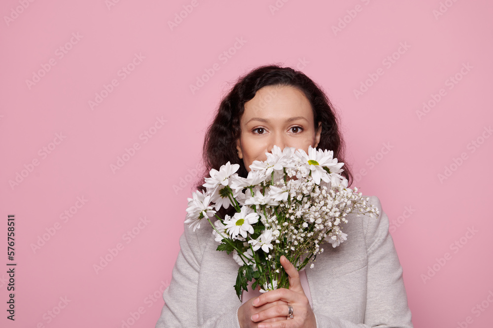 Close-up of a dark-haired middle-aged pretty woman, wearing stylish jacket, looking at camera through a cute bouquet of white spring flowers for women's or Mother's Day, isolated color pink background Close-up of a dark-haired middle-aged pretty woman, wearing stylish jacket, looking at camera through a cute bouquet of white spring flowers for women's or Mother's Day, isolated color pink background