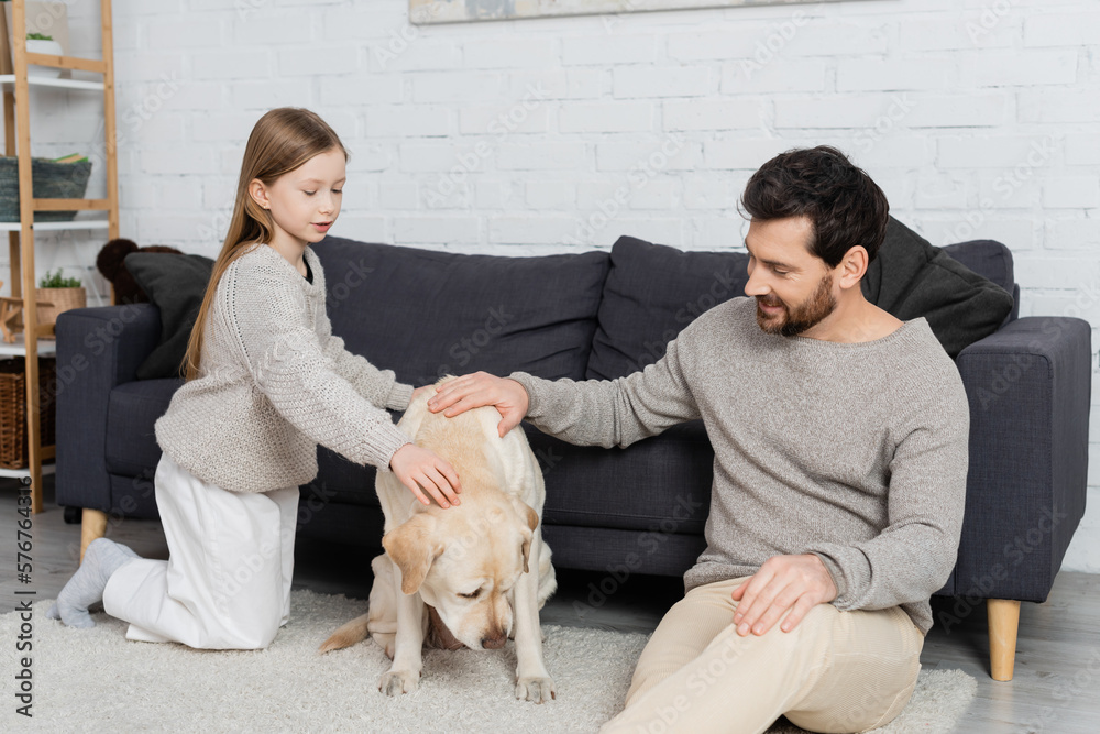 Φωτογραφία happy father and daughter sitting on floor near couch and petting labrador dog
