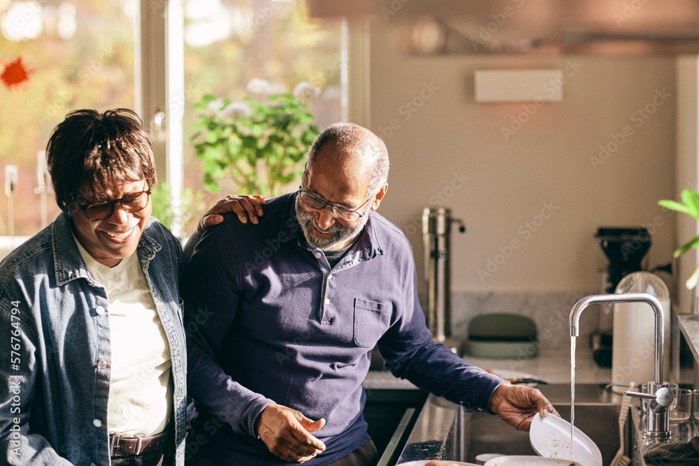 Happy senior woman with hand on shoulder of man washing dishes in kitchen at home Happy senior woman with hand on shoulder of man washing dishes in kitchen at home