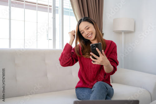 Happy young Asian business woman successful excited raised hand rejoicing with smartphone at home.